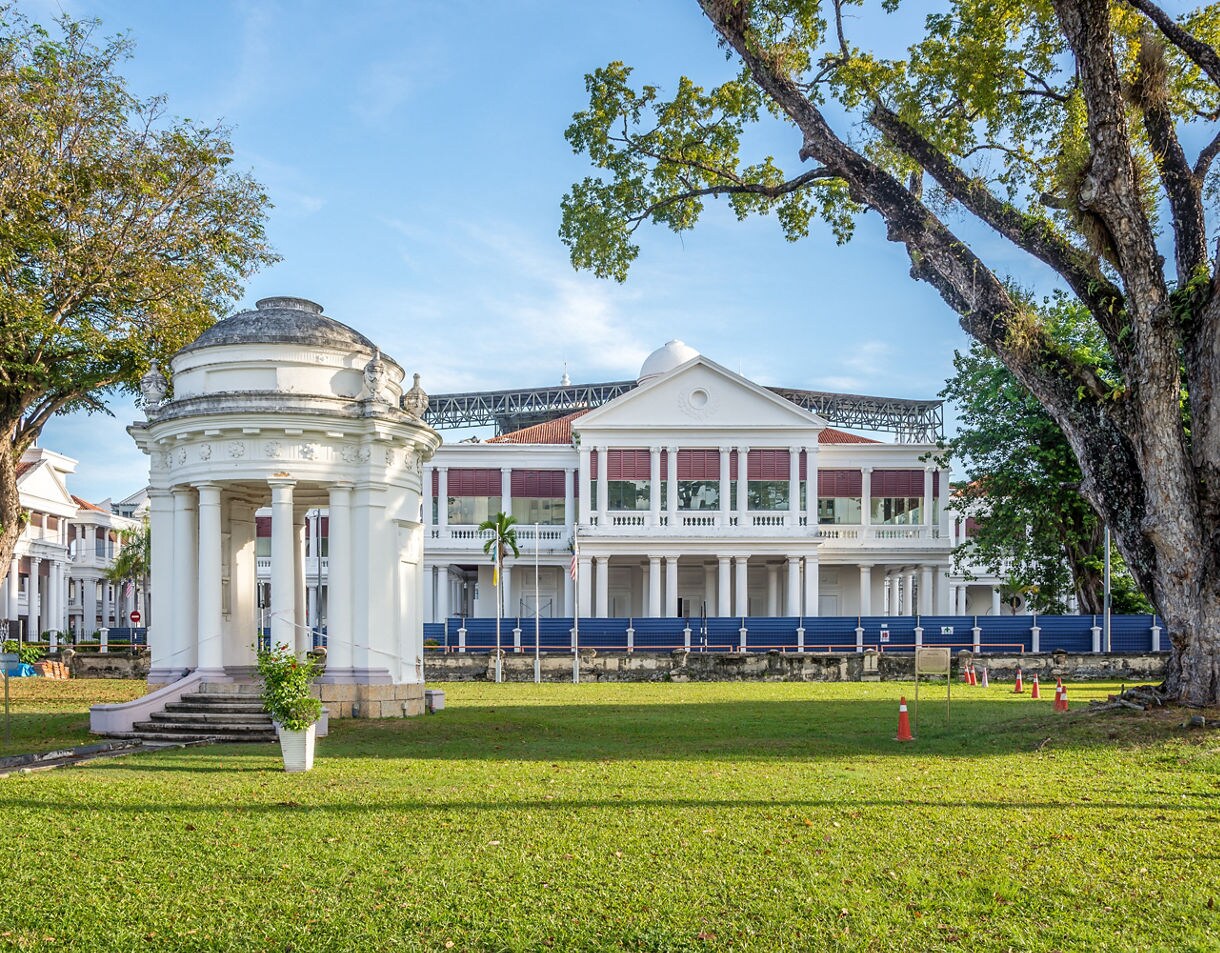White rotunda memorial with columns and dome in front of a colonial-era building with red shutters in George Town, Penang.