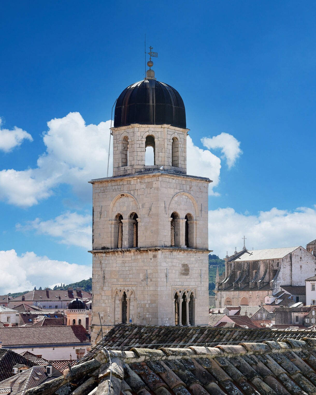 Stone bell tower of the Franciscan Monastery in Dubrovnik, Croatia, with its dark dome and arched openings, standing tall over red-tiled rooftops against a bright blue sky.