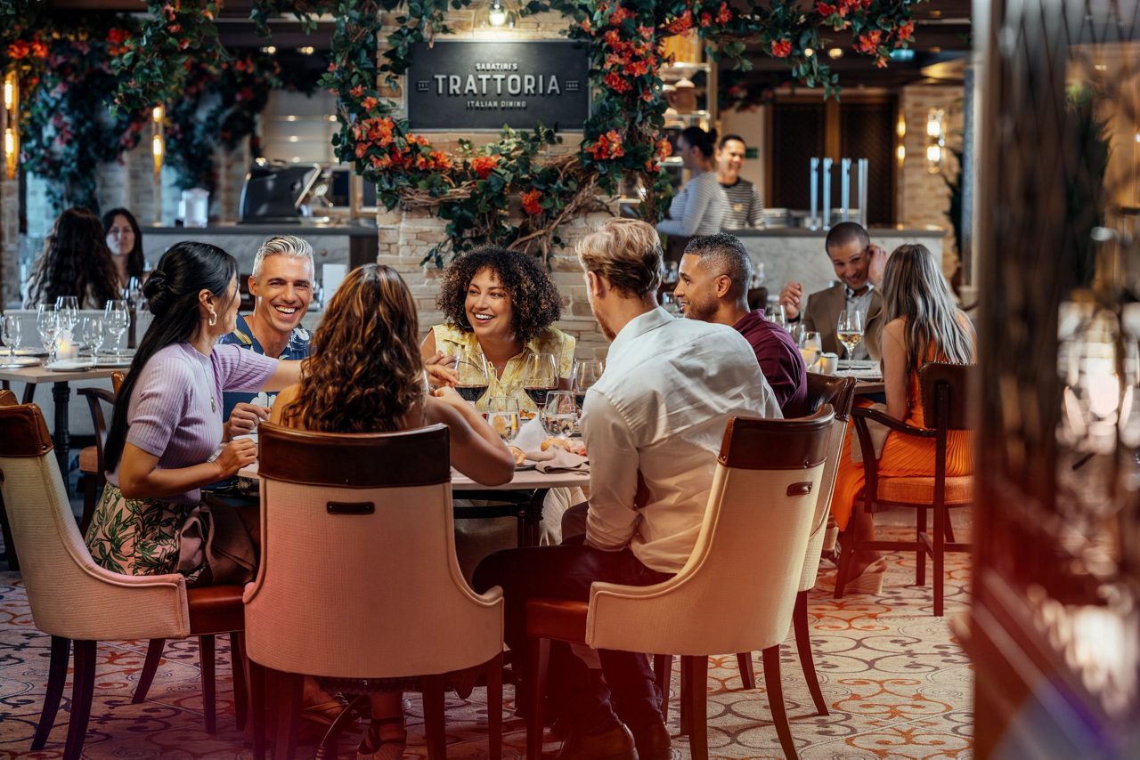 A group of adults sit around a table enjoying a meal in a stylish Italian trattoria. The restaurant interior features warm lighting, floral decorations, and neatly set tables with wine glasses and plates. Diners are engaged in conversation, creating a lively and social atmosphere. A sign reading "TRATTORIA" is visible above the group, emphasizing the Italian dining theme.