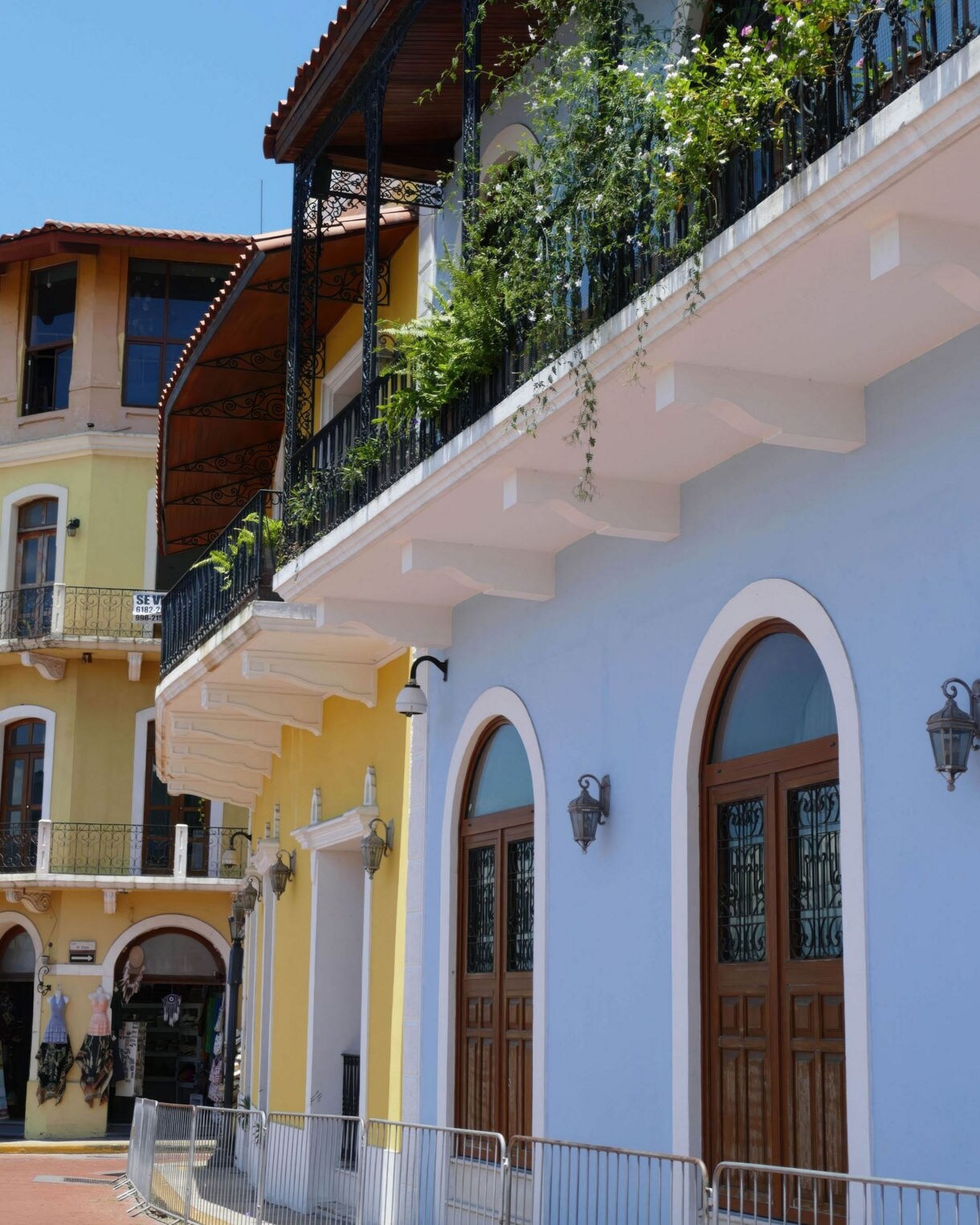 Colorful colonial-style buildings painted in pastel blue and yellow with arched wooden doors, wrought-iron balconies and hanging plants.