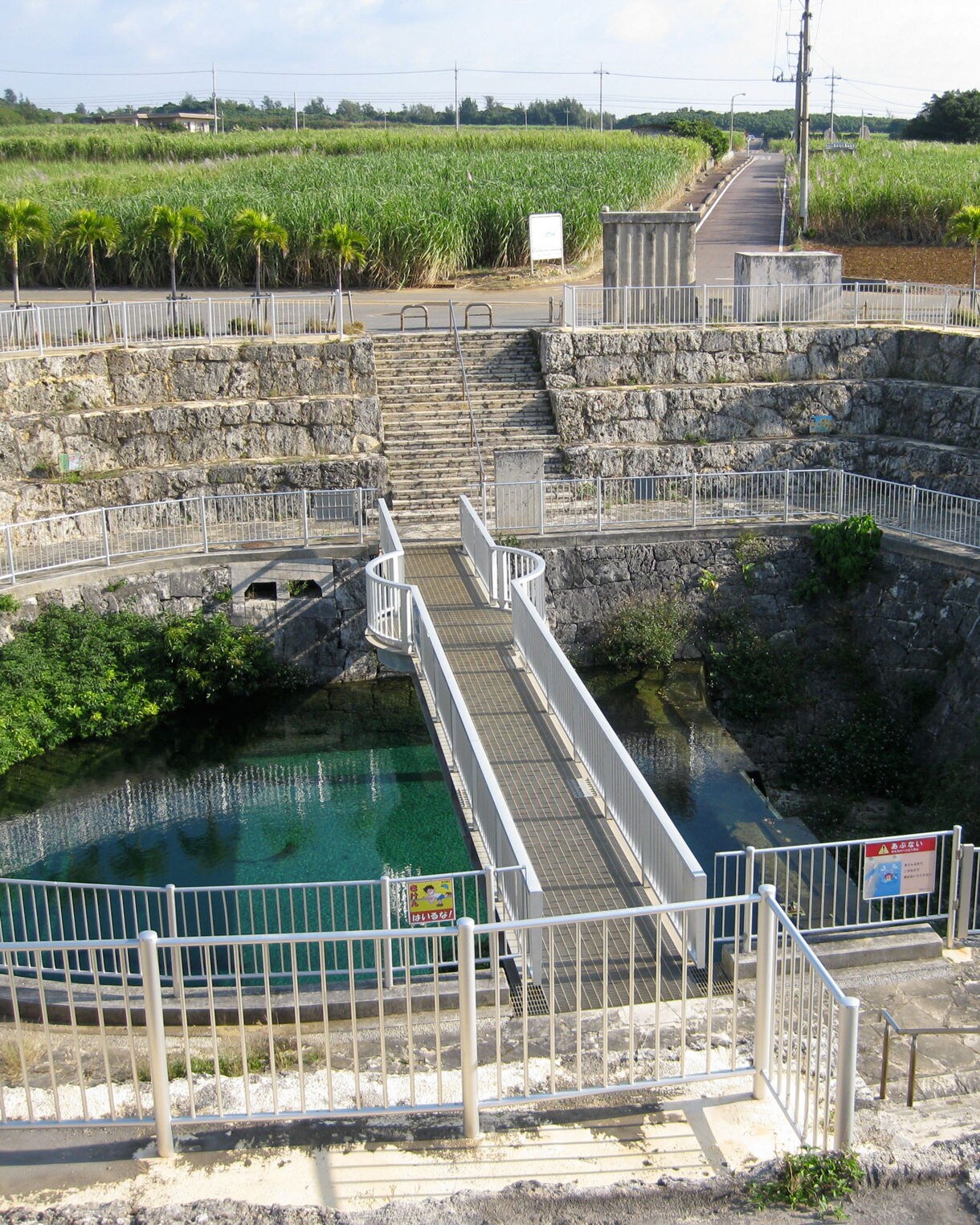 A sunken circular stone basin with a walkway leading over clear turquoise water at the Fukuzato Underground Dam on Miyakojima, surrounded by sugarcane fields and small palm trees under a bright sky.