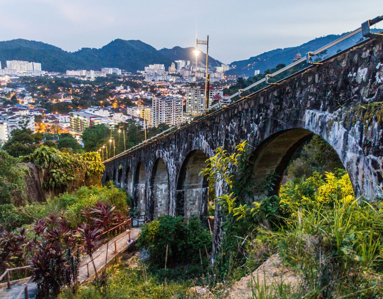 Stone arched funicular bridge on Penang Hill with city lights and high-rises visible in the distance.