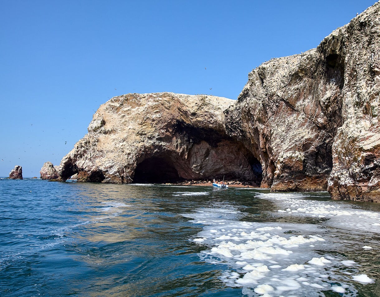Rocky coastal cliffs at the Ballestas Islands with large sea caves, seabirds overhead and a small tour boat approaching the shaded cavern.