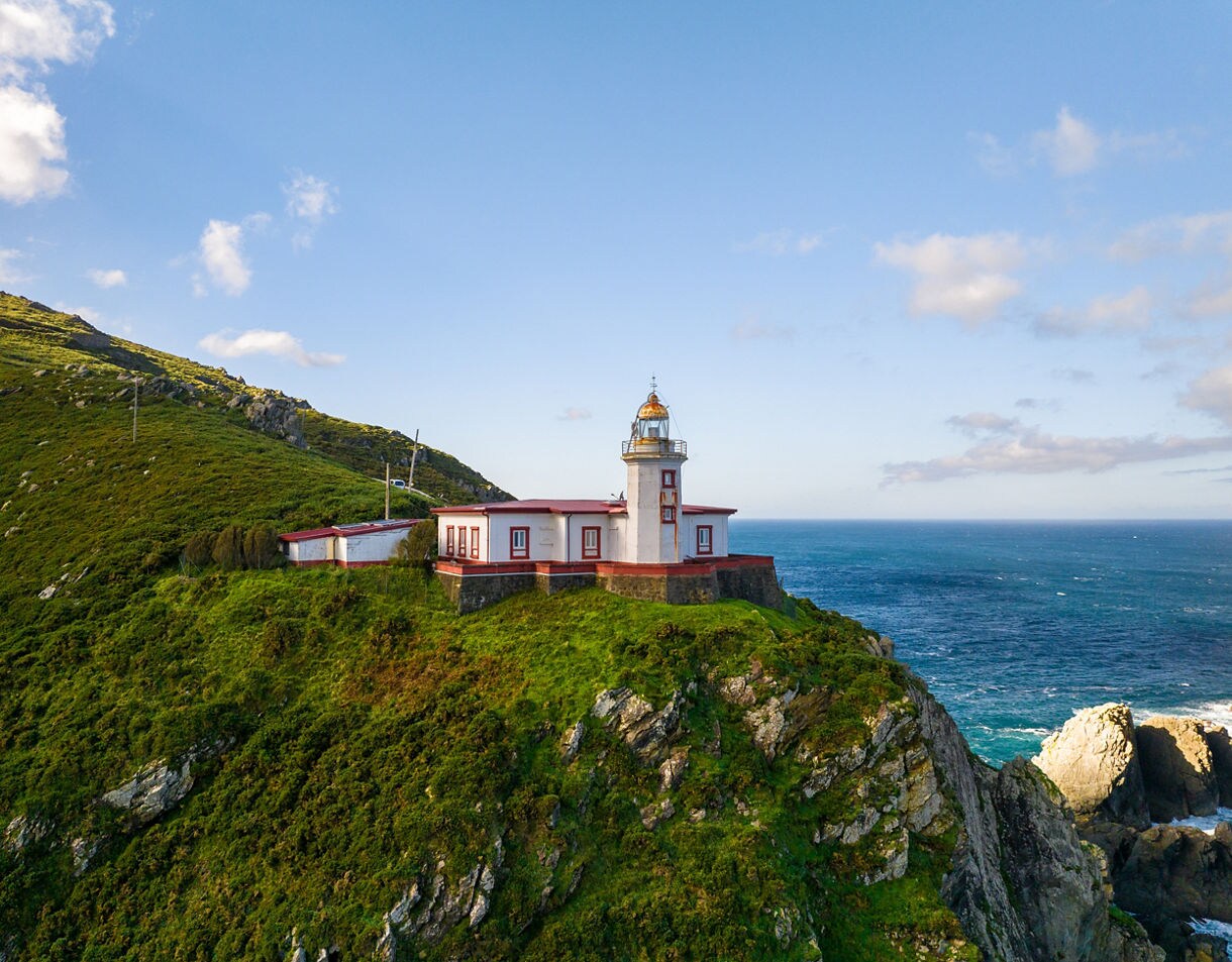 A white and red lighthouse perched on a grassy cliff above the ocean at Cabo Ortegal in Galicia, with rugged rocks below and blue water stretching to the horizon.