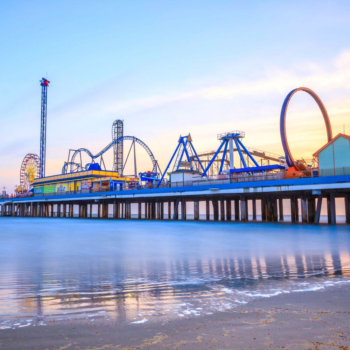 Sunset view of the Galveston Island Historic Pleasure Pier with roller coasters, a  Ferris wheel, and amusement rides over the ocean, reflecting on the water below.