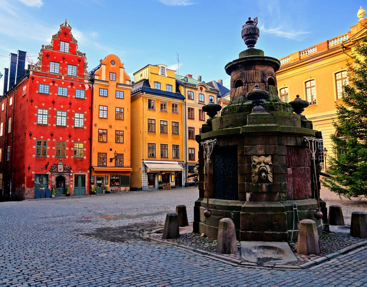 Historic square in Stockholm’s Gamla Stan with cobblestone streets, a stone fountain in the foreground and tall red, yellow and green buildings with decorative facades in the background.