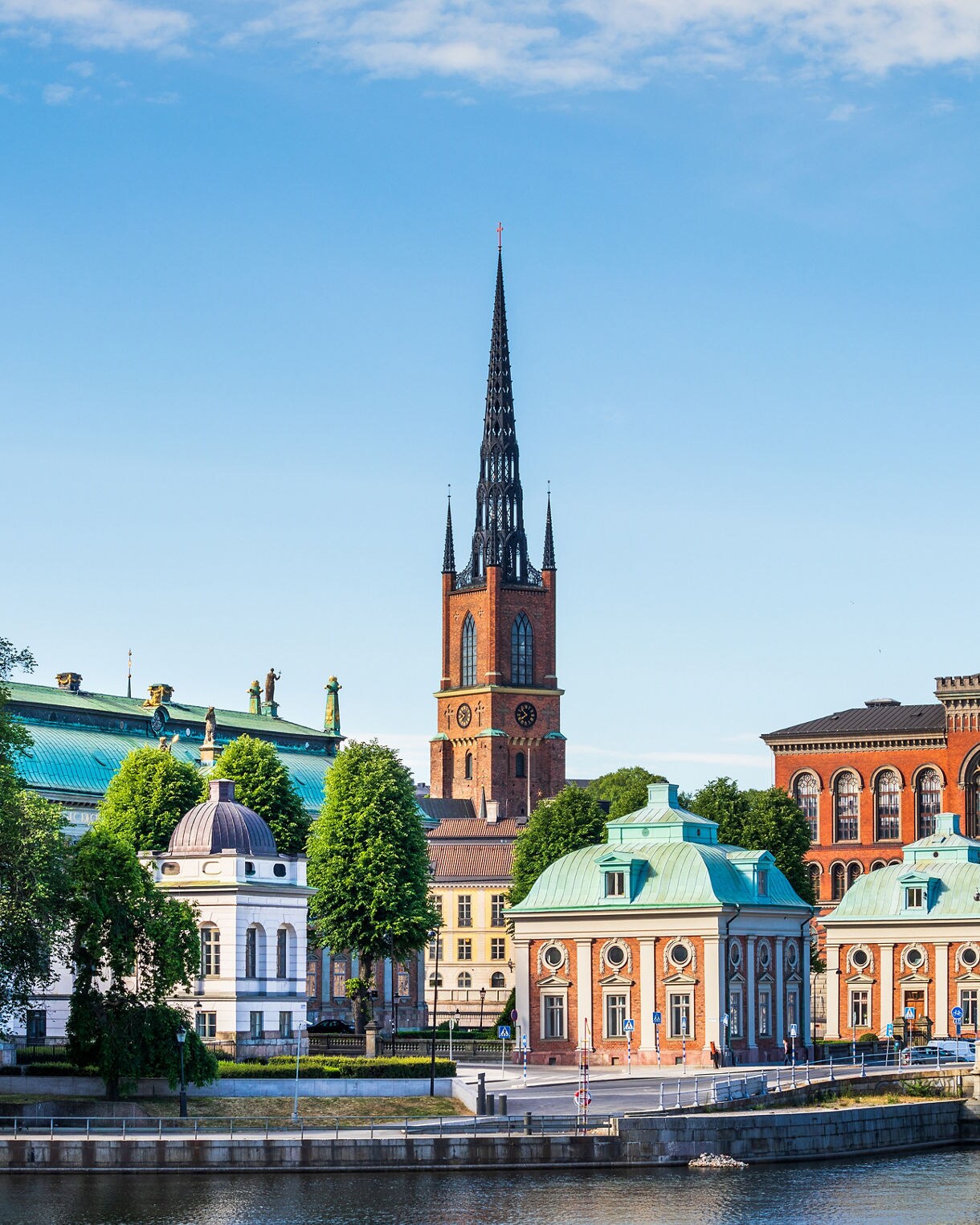 Waterfront view of Riddarholmen Church in Stockholm, with its tall black spire rising behind historic buildings and green copper roofs under a clear sky.