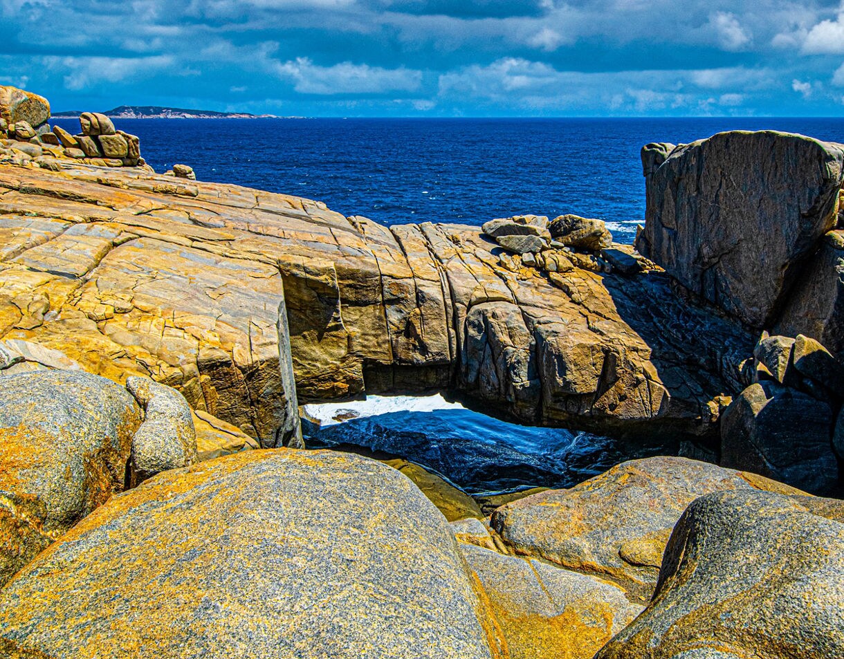 Rugged granite cliffs and boulders form a natural bridge over churning ocean water, with deep blue sea and clouds in the distance.