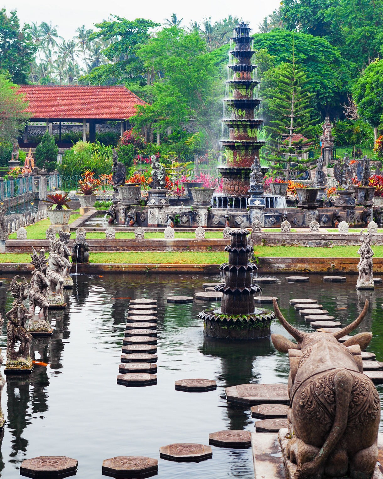 Expansive view of a Balinese water garden featuring tiered fountains, carved stone statues and stepping stones set in reflective pools, surrounded by lush greenery and vibrant tropical plants.