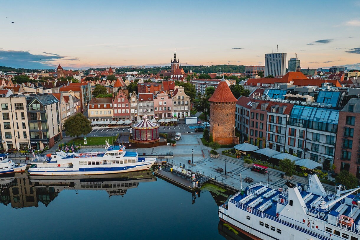 Aerial view of Gdańsk, Poland, showing colorful historic buildings, a round brick tower, moored boats and ferries along the waterfront.