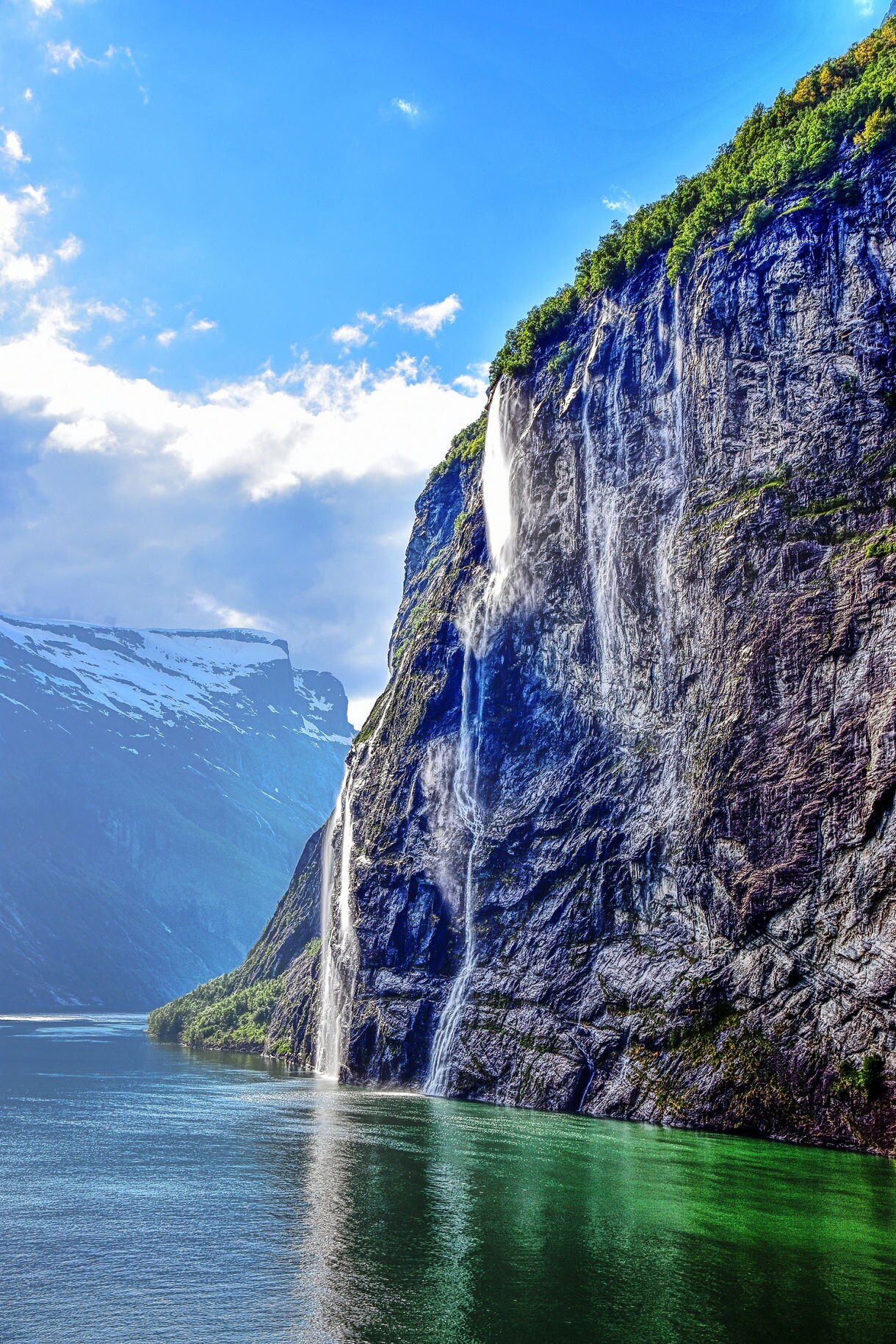 Tall, narrow waterfalls cascade down a steep cliff into the green waters of Norway’s Geirangerfjord under a bright blue sky.