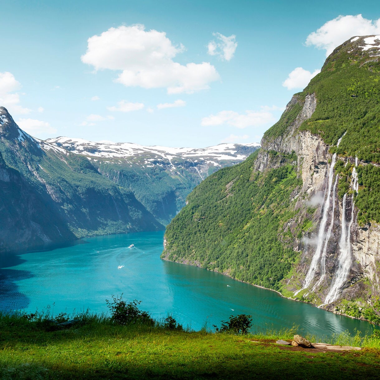 A tall multi-stream waterfall cascades down a steep green cliff into Norway’s turquoise Geirangerfjord, with snow-tipped mountains rising in the distance.