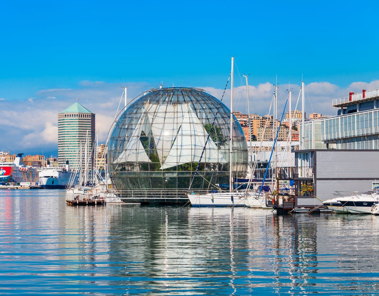 Glass BioSphere and moored sailboats in Genoa’s harbor with city skyscrapers and ferries in the background on a bright day.