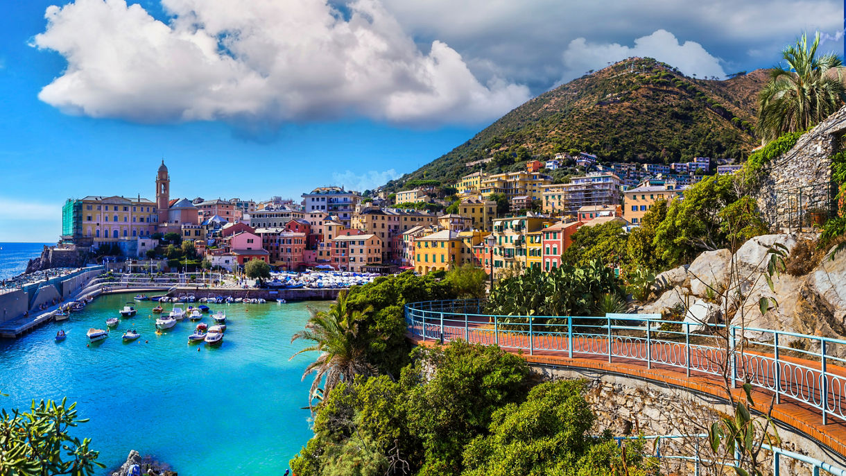 Panoramic view of Genoa, Italy, featuring vibrant waterfront buildings, a small harbor filled with boats and lush hills rising behind the city.