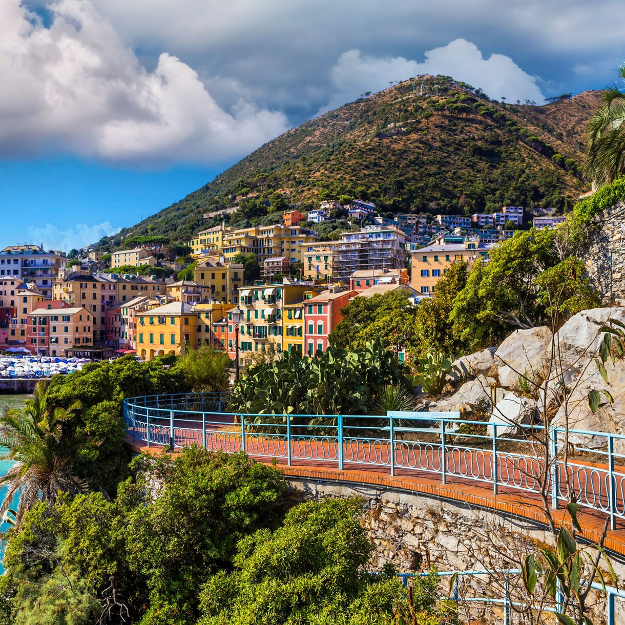  Panoramic view of Genoa, Italy, featuring vibrant waterfront buildings, a small harbor filled with boats and lush hills rising behind the city.