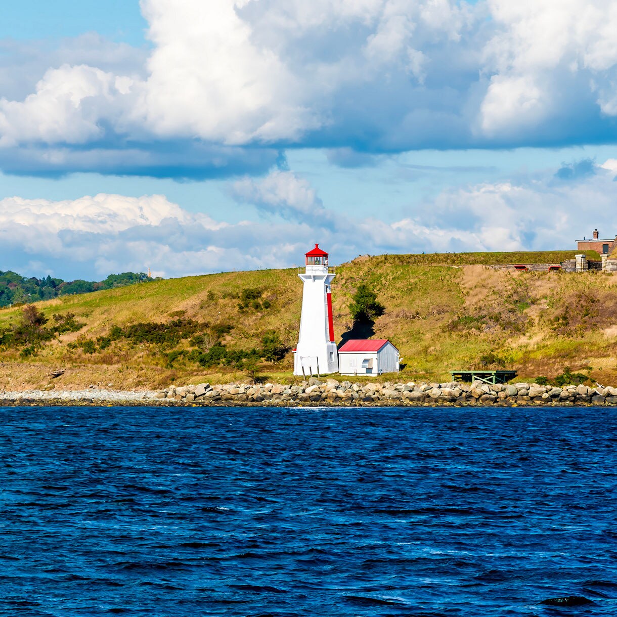 White lighthouse with a red roof on Georges Island, set on grassy slopes above rocky shoreline with deep blue water in the foreground.