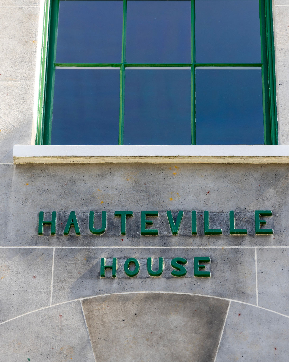 A close-up view shows green lettering spelling "HAUTEVILLE HOUSE" set against a light grey stone wall beneath a green-framed window.