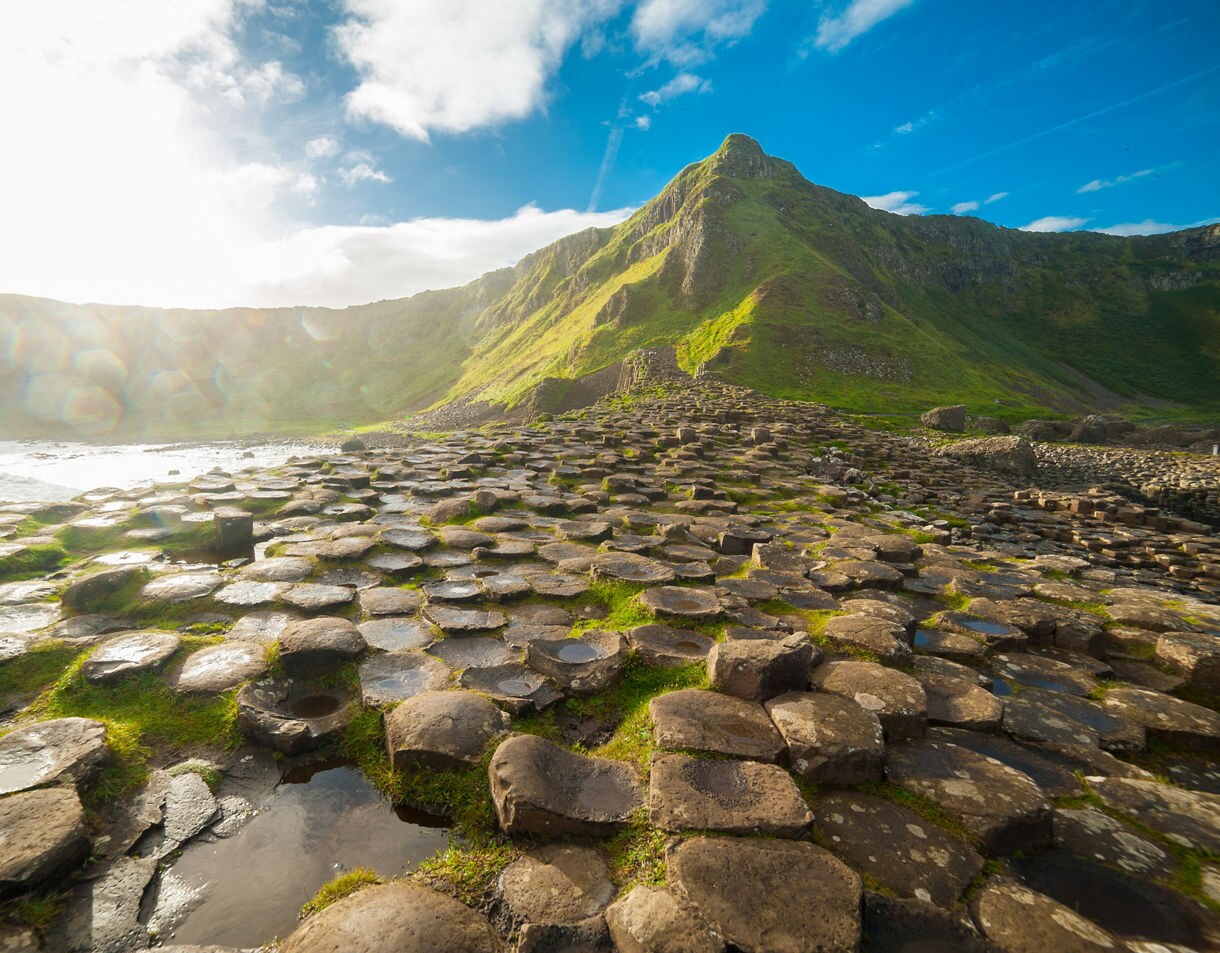 Hexagonal basalt columns of the Giant’s Causeway in Northern Ireland with sunlight reflecting off the sea and green hills rising in the background.