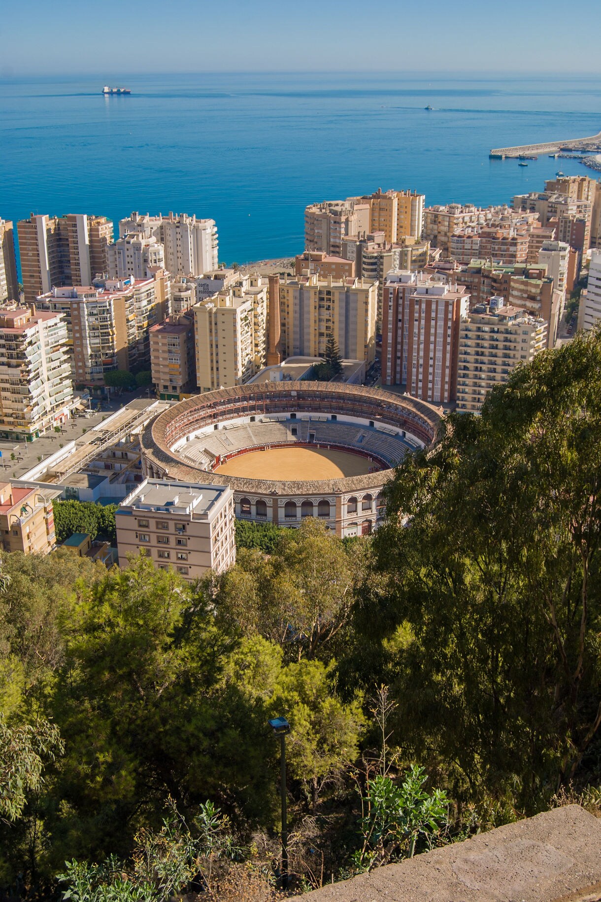Aerial view of Málaga’s bullring surrounded by tall apartment buildings, with the Mediterranean Sea in the background.