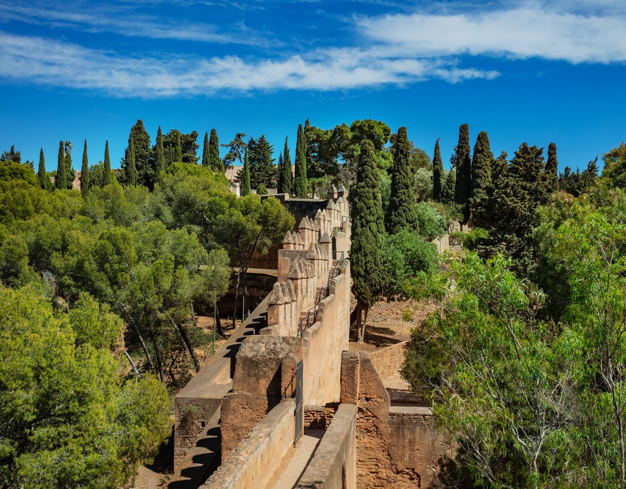 Stone battlements of Gibralfaro Castle in Málaga, Spain, surrounded by dense green trees under a bright blue sky.