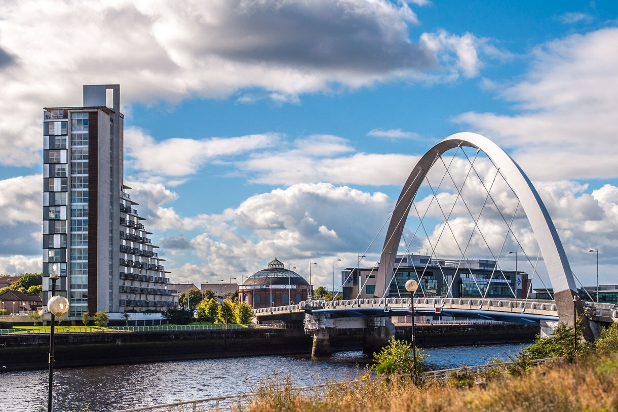 The Squinty Bridge over the River Clyde in Glasgow with modern high-rise buildings and a partly cloudy sky.