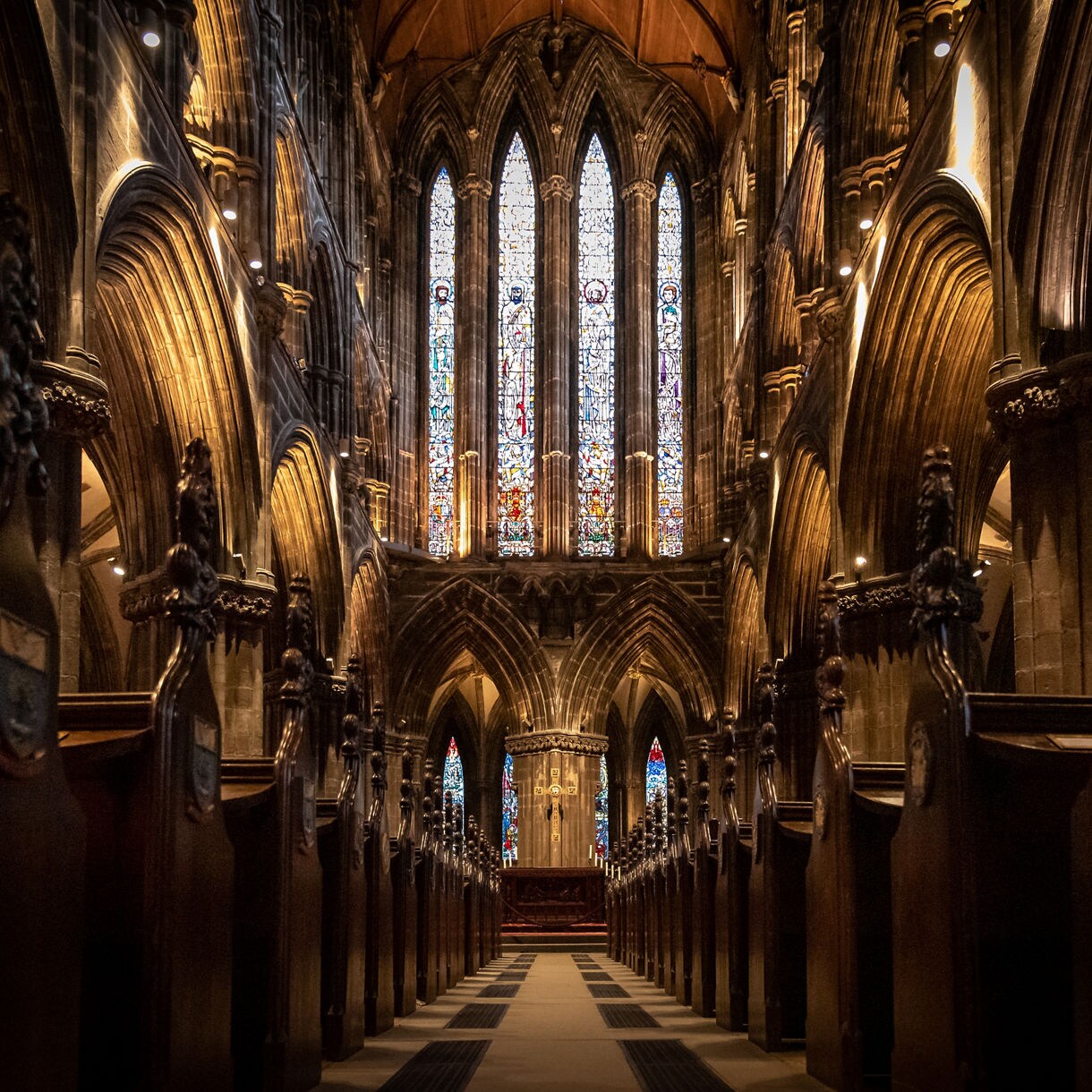Interior view of Glasgow Cathedral with tall Gothic arches, rows of dark wooden pews and a large stained glass window glowing at the far end.