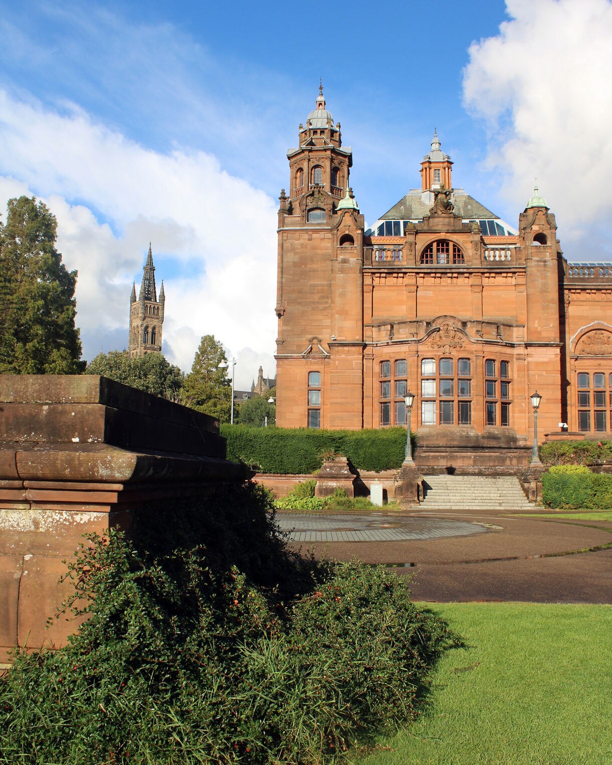 Kelvingrove Art Gallery and Museum in Glasgow, a grand red sandstone building with ornate towers, set against a bright blue sky with greenery in the foreground.