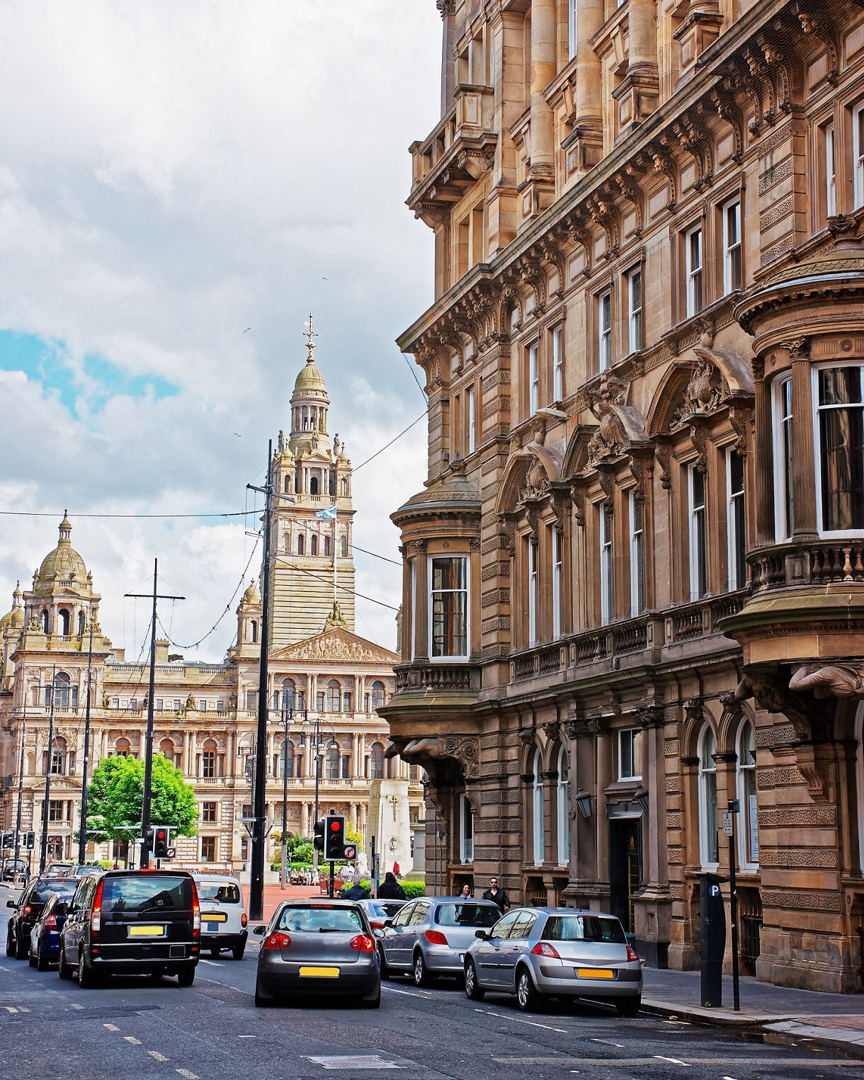 Street view in Glasgow with rows of historic sandstone buildings, cars lining the road and the grand towers of the City Chambers rising in the background under a partly cloudy sky.