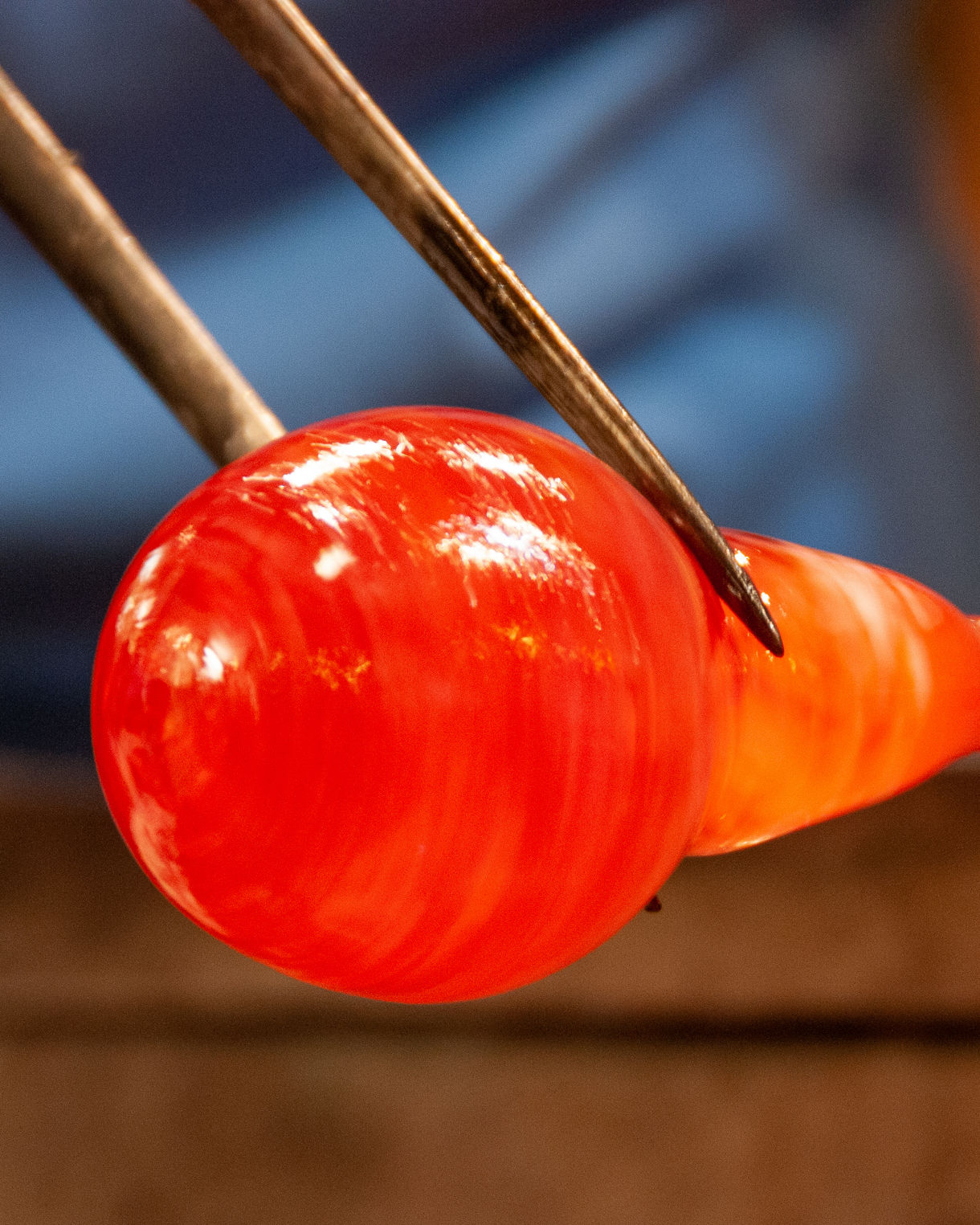Close-up of a glassblower shaping a glowing orange sphere of molten glass with metal tools.