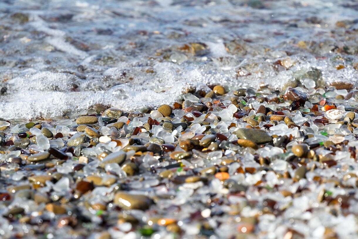 A close-up view of Glass Beach in Fort Bragg, California, featuring smooth, multicolored sea glass and pebbles along the shoreline as ocean waves gently wash over them.