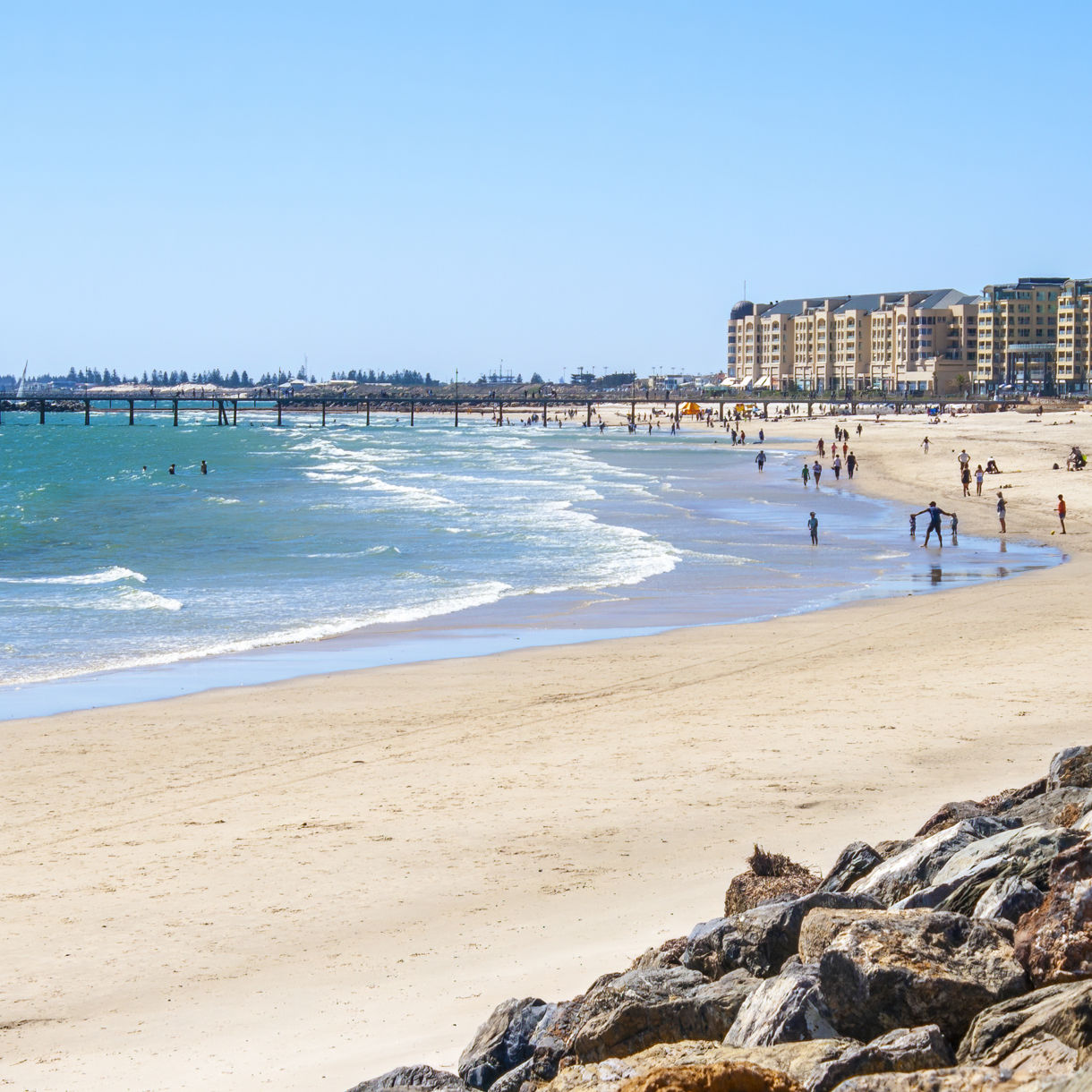 People walking and swimming along Glenelg Beach in Adelaide, with a long pier extending into the turquoise water and beachfront buildings in the distance.