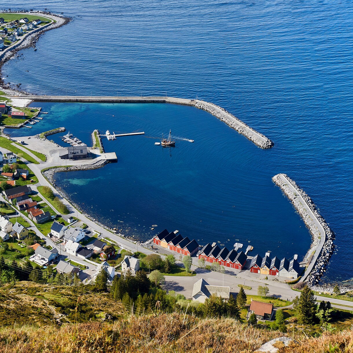 Aerial view of Godøy, Norway, showing a small coastal village with colorful houses, a lighthouse and a sheltered harbor beside the open ocean.