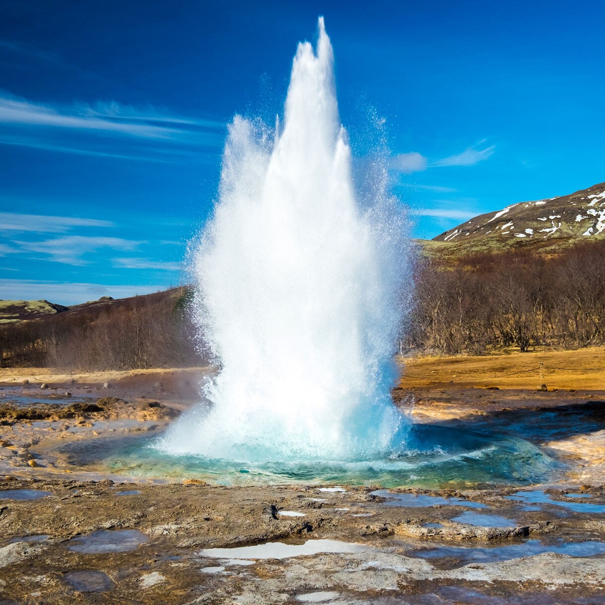 Strokkur geyser in Iceland erupting with a powerful jet of water and steam, surrounded by rocky ground and distant snow-dusted hills under a bright sky.