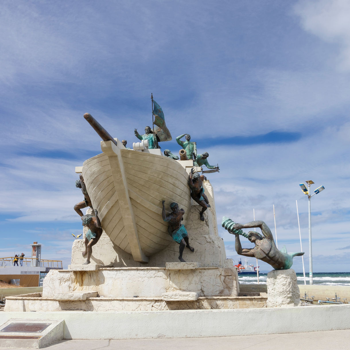 Monument featuring a large stone ship surrounded by dynamic bronze figures near the waterfront in Punta Arenas, set against a bright blue sky.