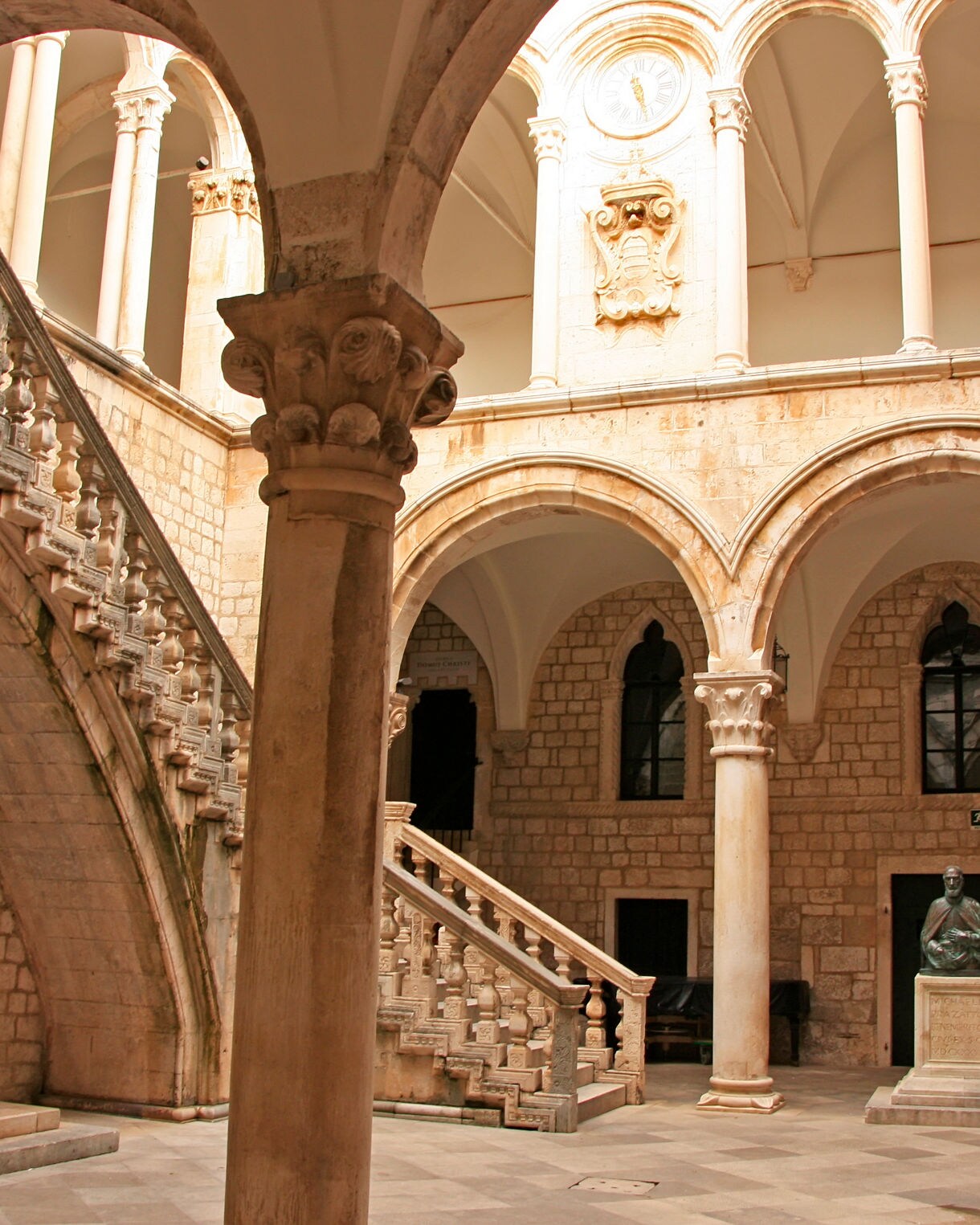 Stone courtyard of Dubrovnik’s Rector’s Palace with arched columns, a grand staircase and a bronze bust statue beneath vaulted ceilings.