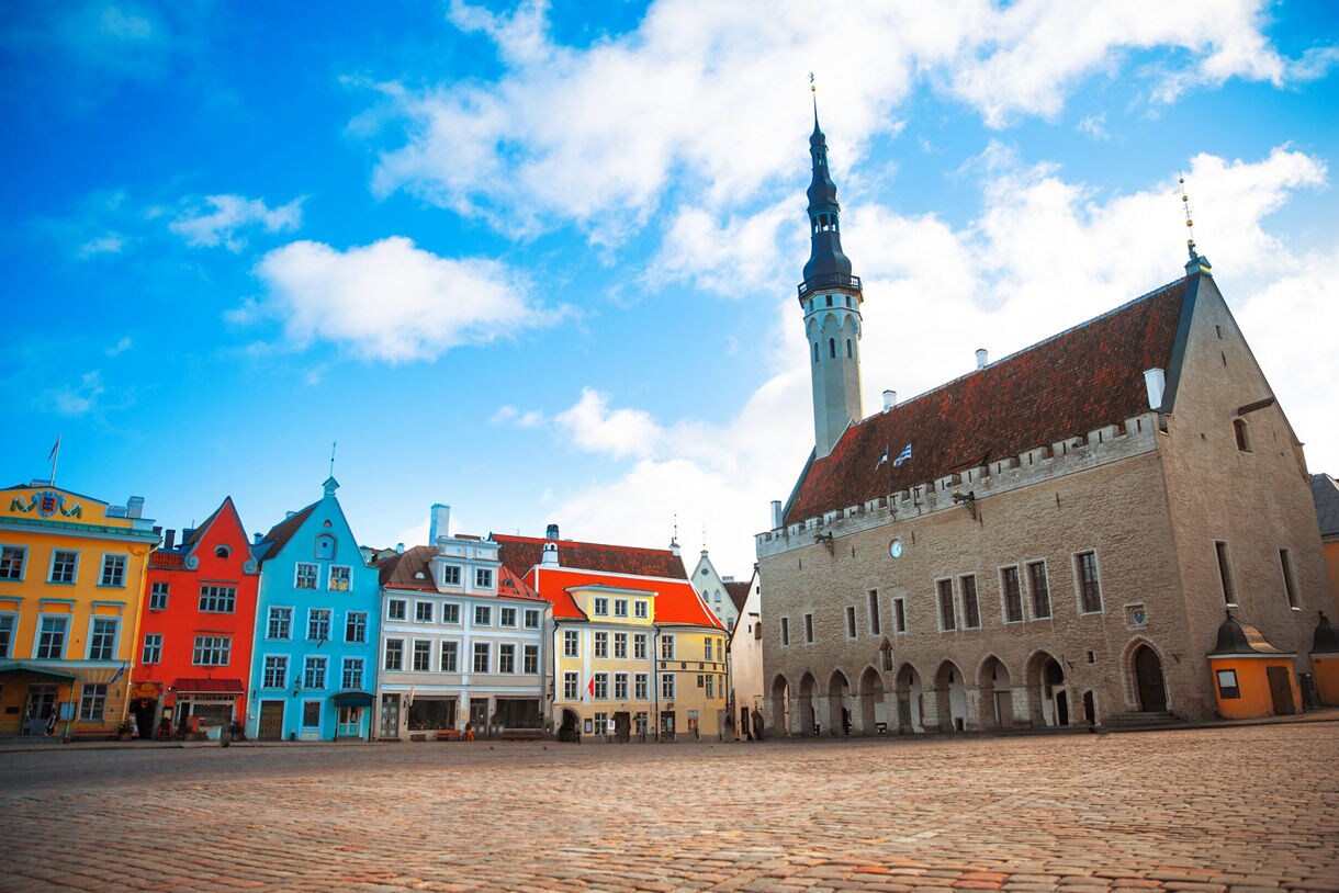 Tallinn Town Hall Square in Estonia, featuring pastel-colored medieval buildings and the Gothic Town Hall with its tall spire rising into a bright blue sky