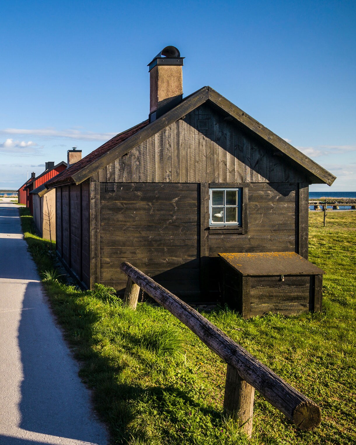 Traditional fishing huts in Gnisvärd, Gotland, Sweden, painted in red and black wood, lined along a narrow road leading toward the coastline.