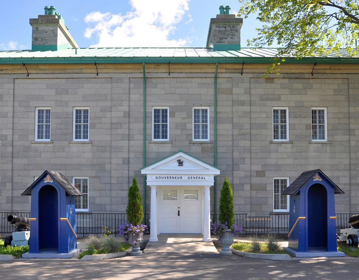 Front view of the Governor’s House in Québec City, showing a stone façade, white entrance with columns, blue guard booths and cannons on either side.