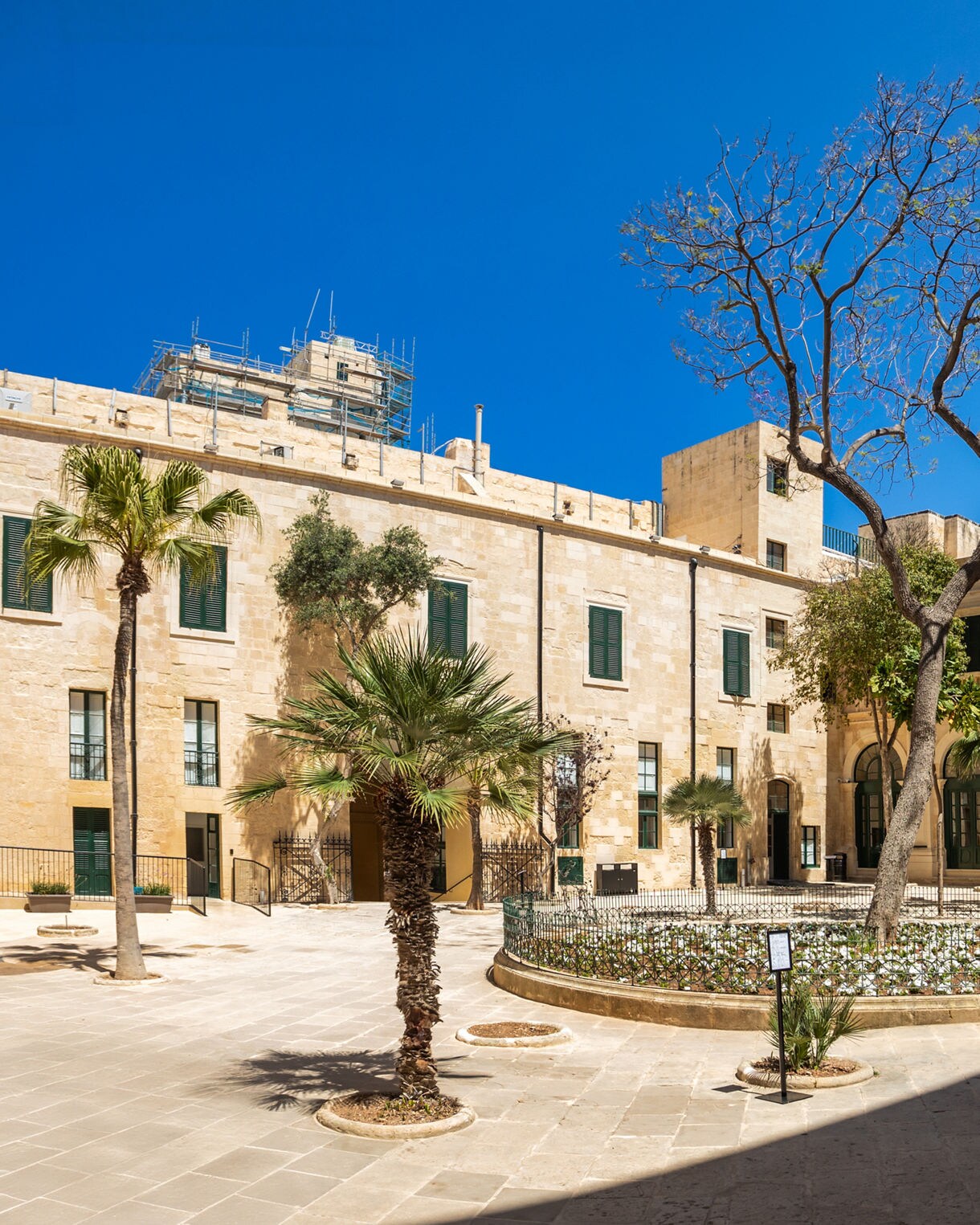 Courtyard of the Grandmaster’s Palace in Valletta, Malta, featuring a central garden with palm trees, a fountain and limestone Baroque architecture under a clear blue sky.