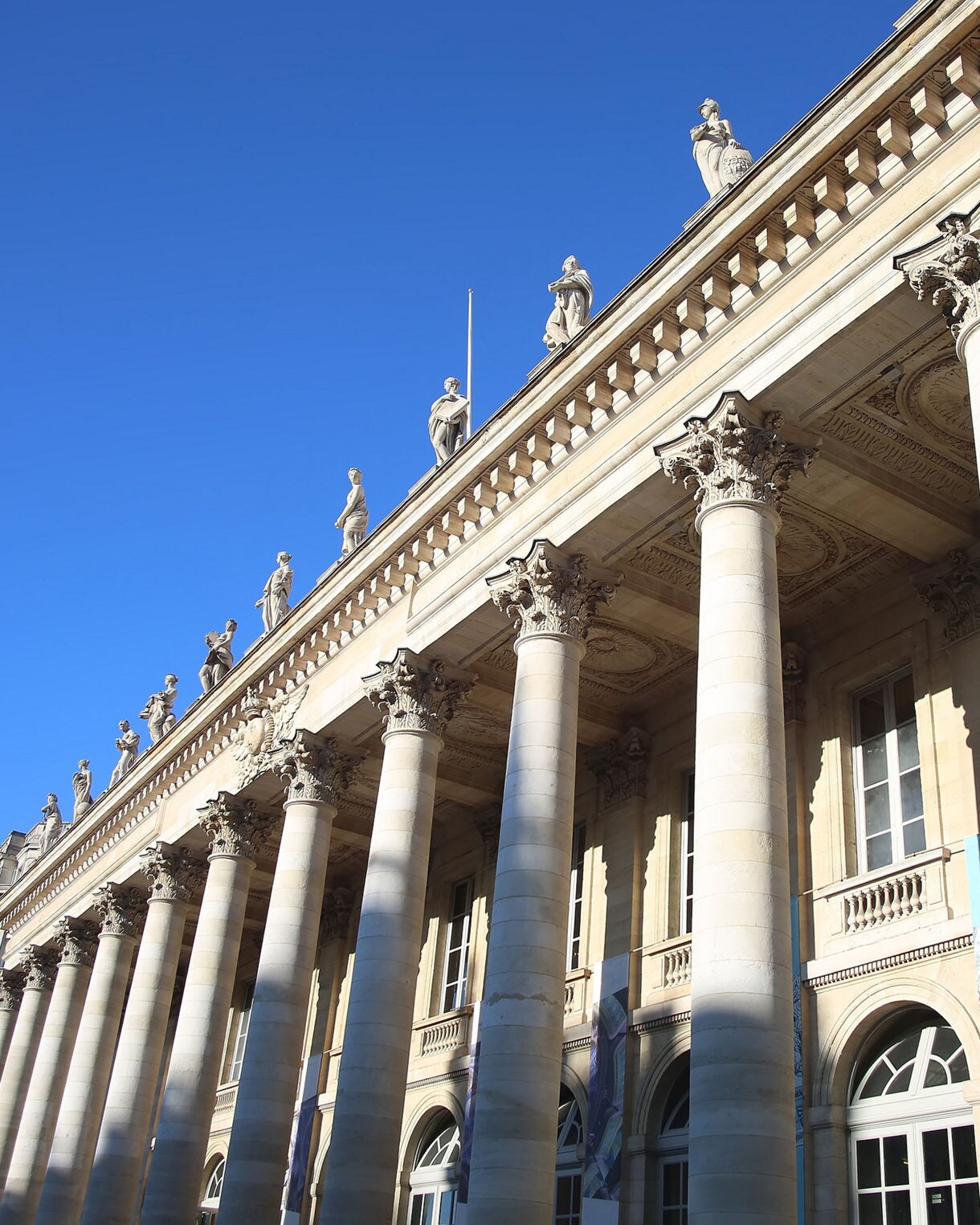 The Grand Théâtre in Bordeaux, featuring tall Corinthian columns and statues along the roofline, illuminated by bright sunlight under a clear blue sky.