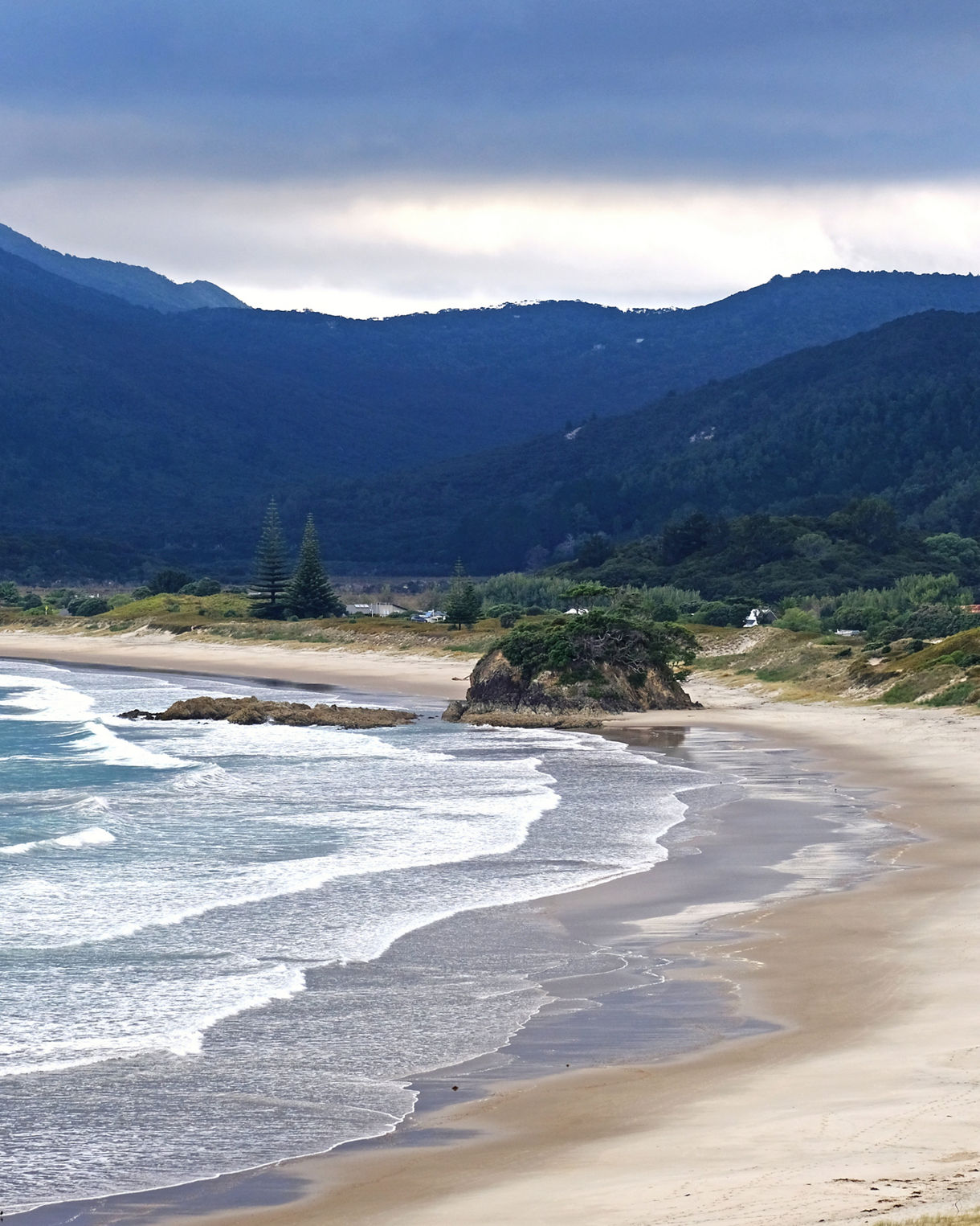 Remote sandy beach on Great Barrier Island with rolling waves, rocky outcrops and dense green mountains in the background.