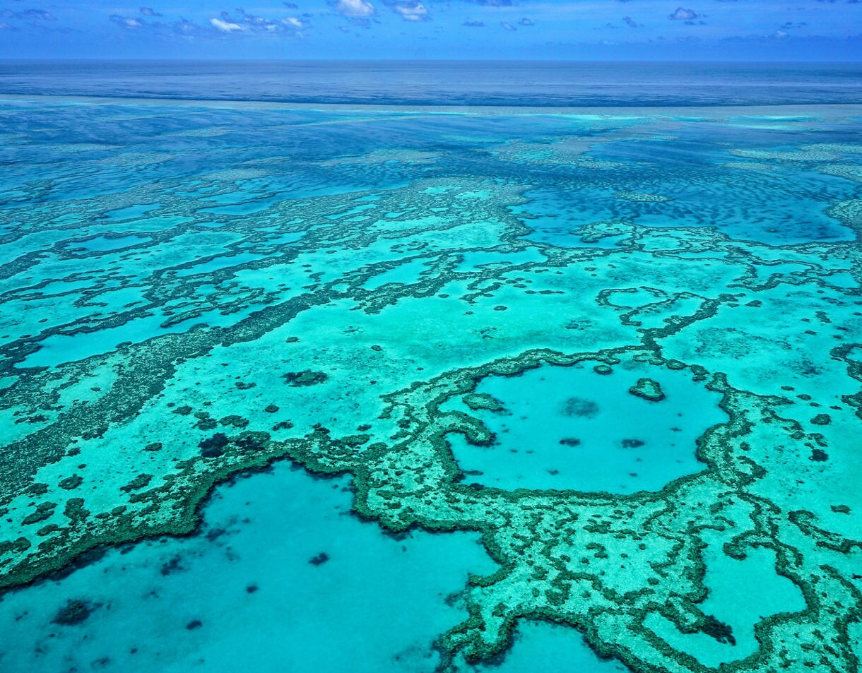 Aerial view of bright turquoise water with intricate coral formations creating swirling patterns across a shallow reef.