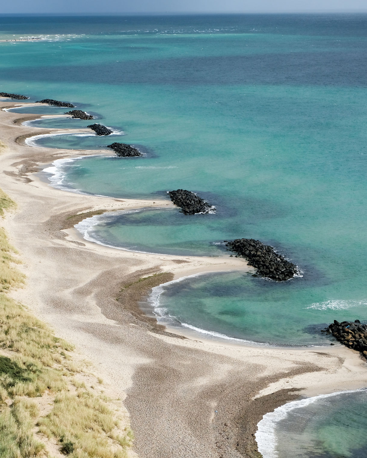 Aerial view of Grenen Beach with curving sandy shoreline, turquoise waters and rock groynes extending into the sea near Skagen.