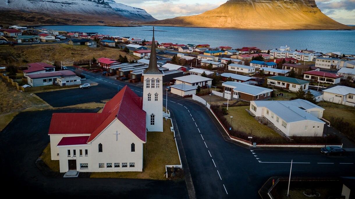 A coastal town with colorful houses and a white church with a tall steeple, set beside a calm bay and framed by snow-dusted mountains and the distinct peak of Kirkjufell glowing in warm sunlight.