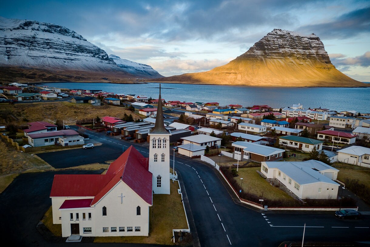 A coastal town with colorful houses and a white church with a tall steeple, set beside a calm bay and framed by snow-dusted mountains and the distinct peak of Kirkjufell glowing in warm sunlight.