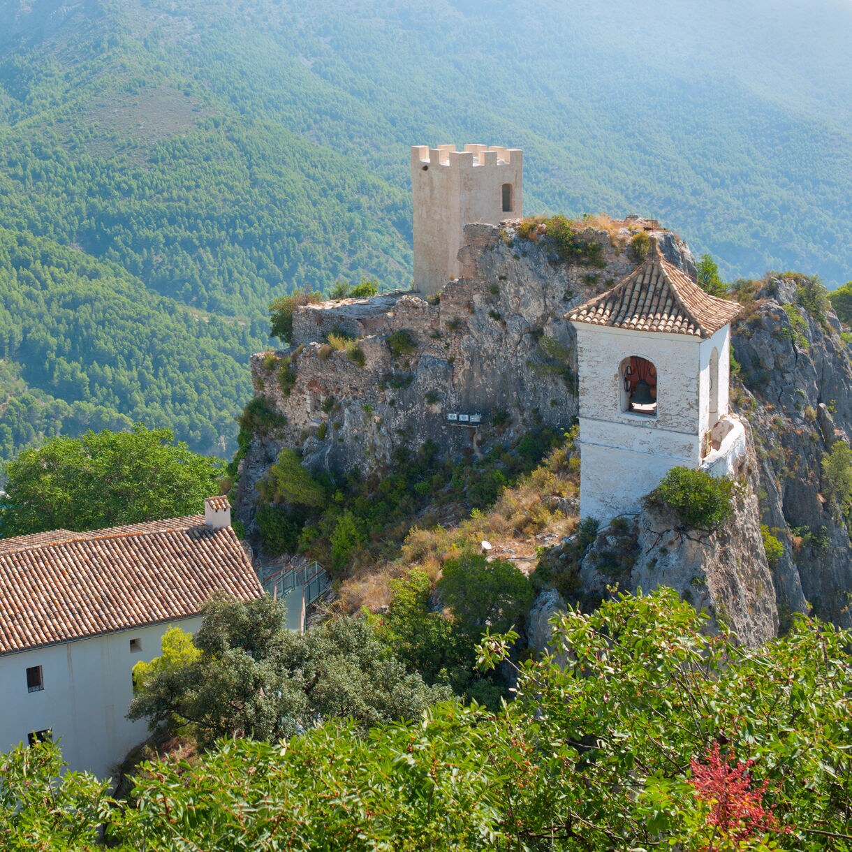 Cliffside view of Guadalest in Spain, featuring a white bell tower perched on a rocky outcrop above a turquoise reservoir, surrounded by forested mountains and traditional terracotta-roofed buildings.