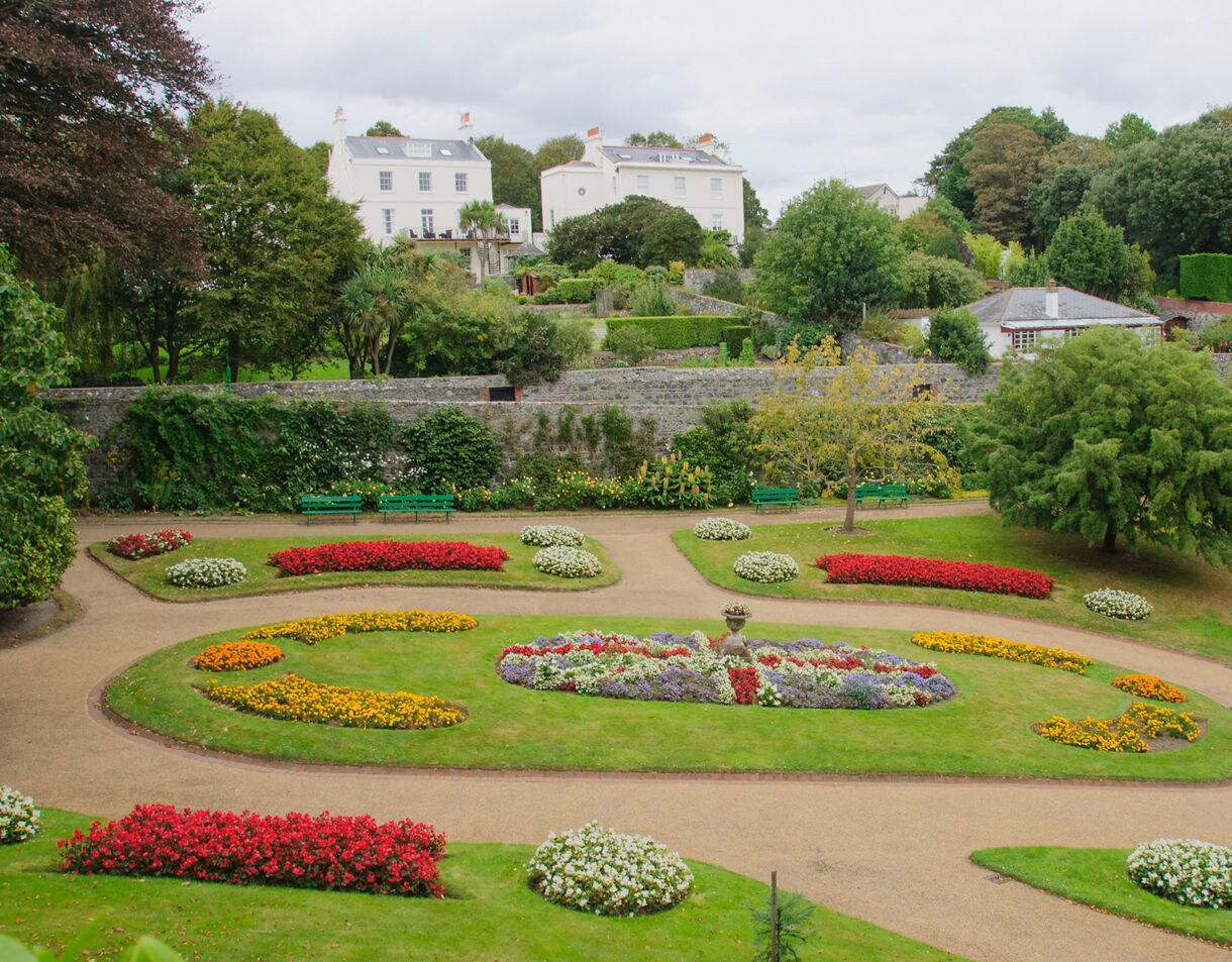 A high-angle view shows neatly manicured Victorian public gardens featuring vibrant circular flower beds, gravel walking paths, green lawn, and white historic buildings nestled among trees in the background under an overcast sky.