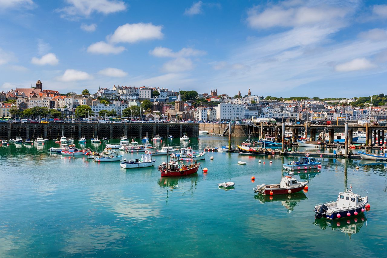 Numerous small fishing and pleasure boats float in a clear turquoise harbor in front of a hillside town filled with historic white and brick buildings beneath a blue sky.