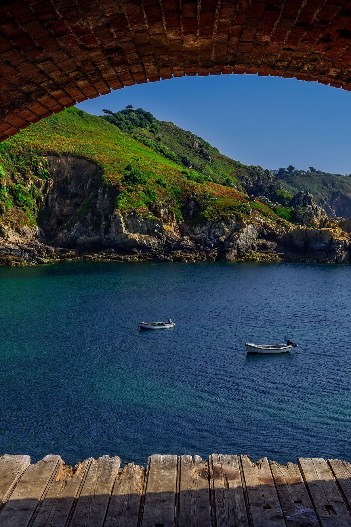 Framed through a red brick archway and wooden floor, a scenic view reveals small boats anchored in clear blue cove waters backed by green, rocky coastal cliffs under a bright sky.