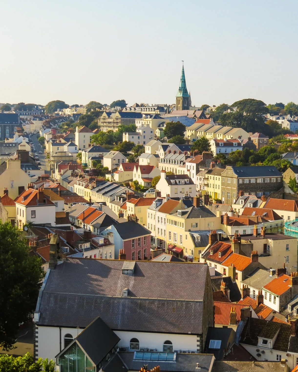 An elevated view shows a prominent stone church standing above red-tiled rooftops in a coastal town overlooking a harbor filled with sailboats under a clear blue sky.